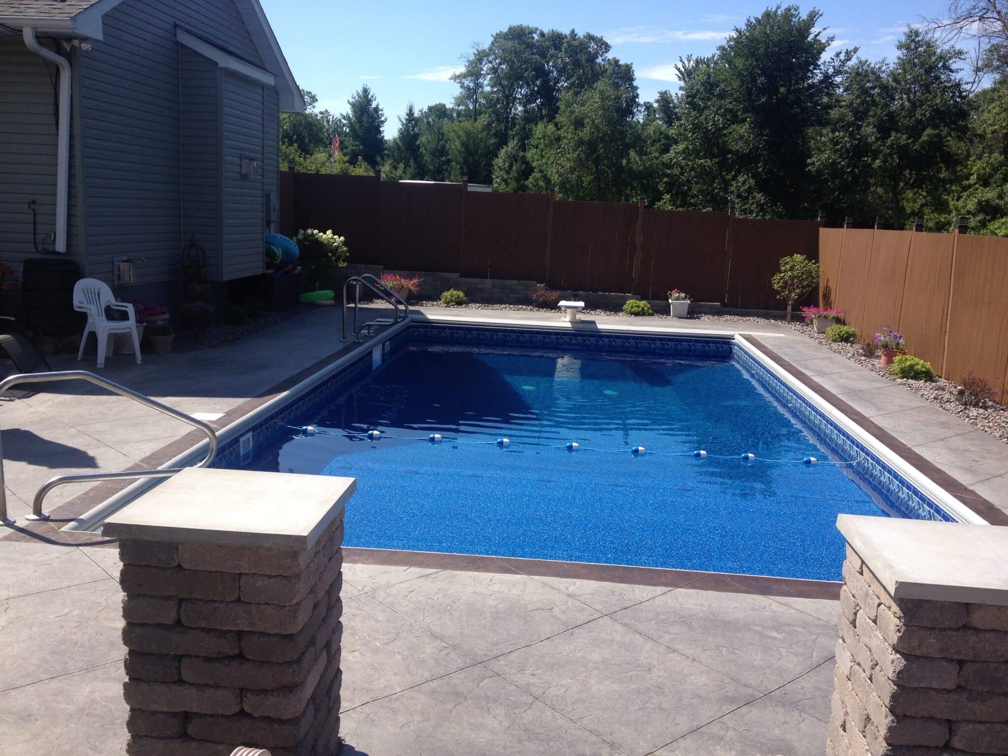 A backyard swimming pool with trees in the background behind a fence.