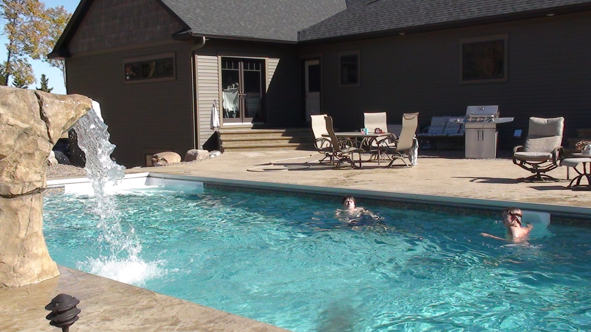 A backyard scene featuring an inground swimming pool with a rock waterfall on the left. The pool has a concrete deck surround where an outdoor dining set and a grill are located. Two people are visible in the pool. The house, with a dark gray roof and siding, is in the background.