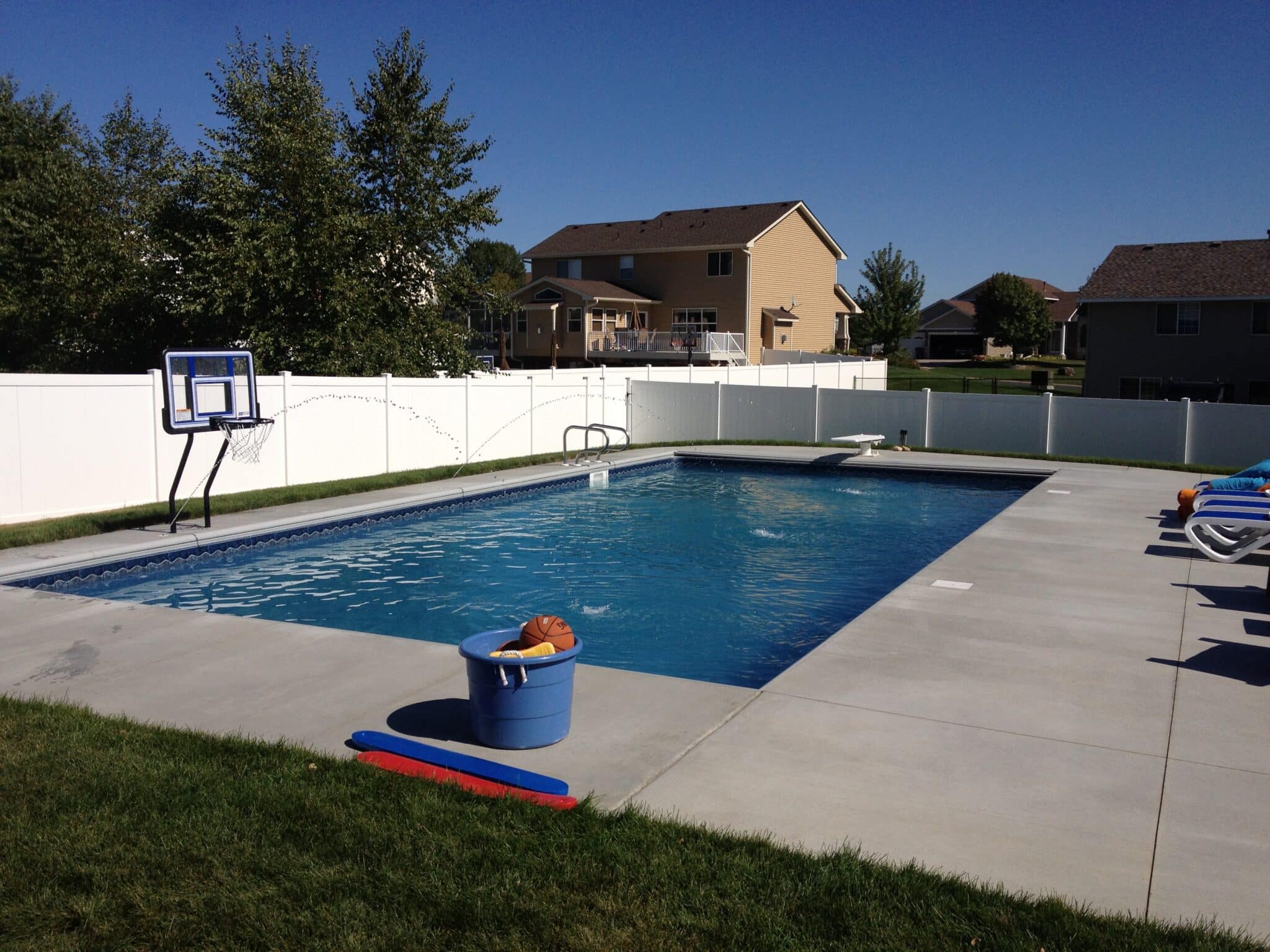 A backyard with an inground swimming pool, a concrete patio, and a white fence. A basketball hoop is at one end of the pool. A blue bucket with pool toys sits on the patio. A two-story house with a beige exterior is visible in the background, and there are trees and a blue sky above.