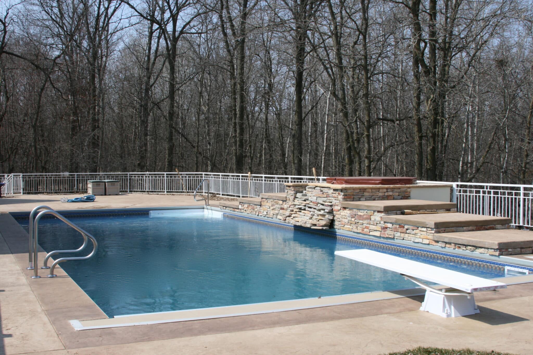 An outdoor swimming pool and a raised hot tub surrounded by a stone wall, with a diving board and a white fence in the background of bare trees.
