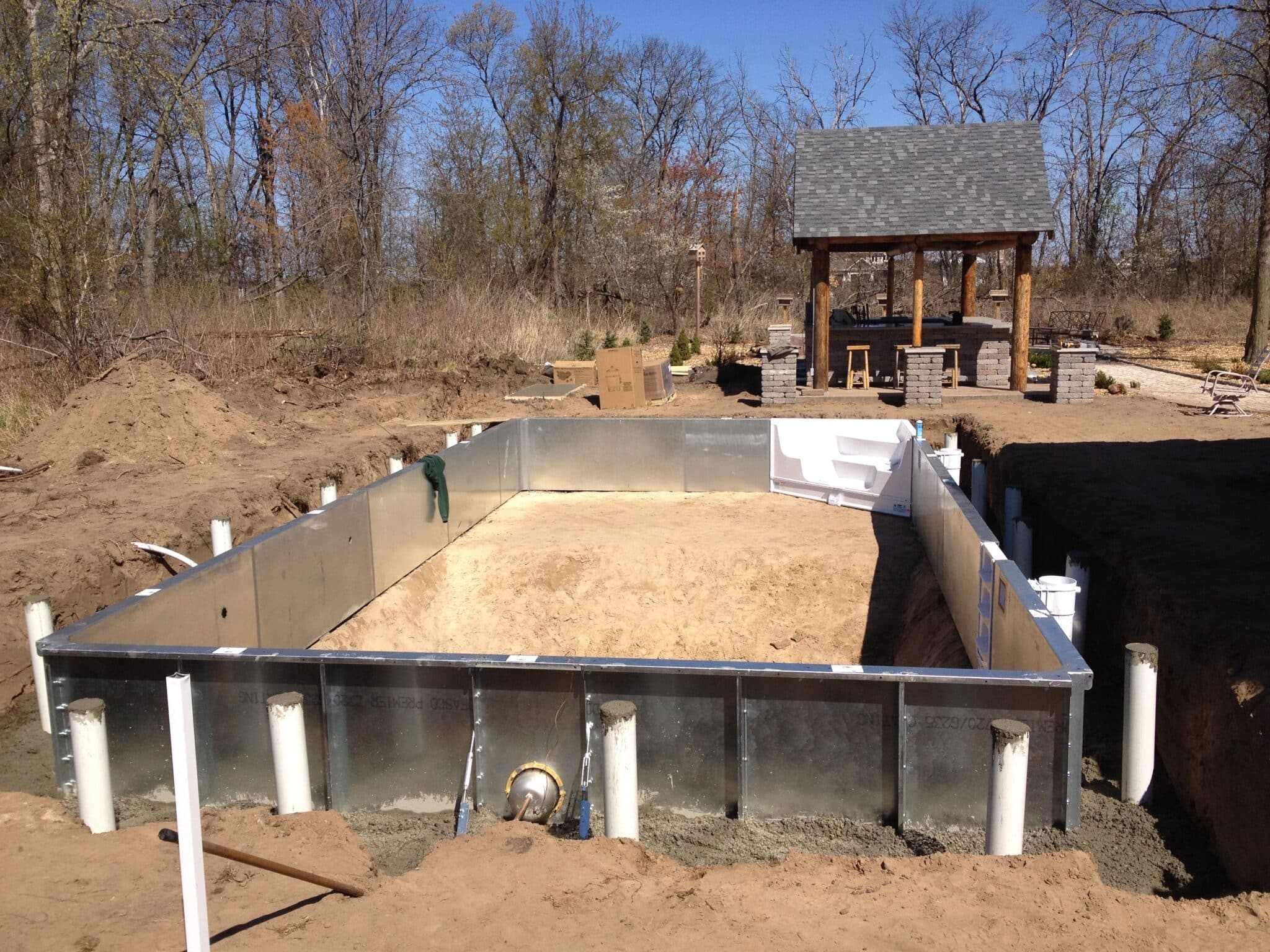 An inground swimming pool under construction, featuring a rectangular perimeter of galvanized steel panels secured in an excavated pit. White PVC support pipes and concrete footings line the exterior walls, while the interior floor is prepared with a layer of sand. In the background, a completed outdoor pavilion and stone bar area are visible.