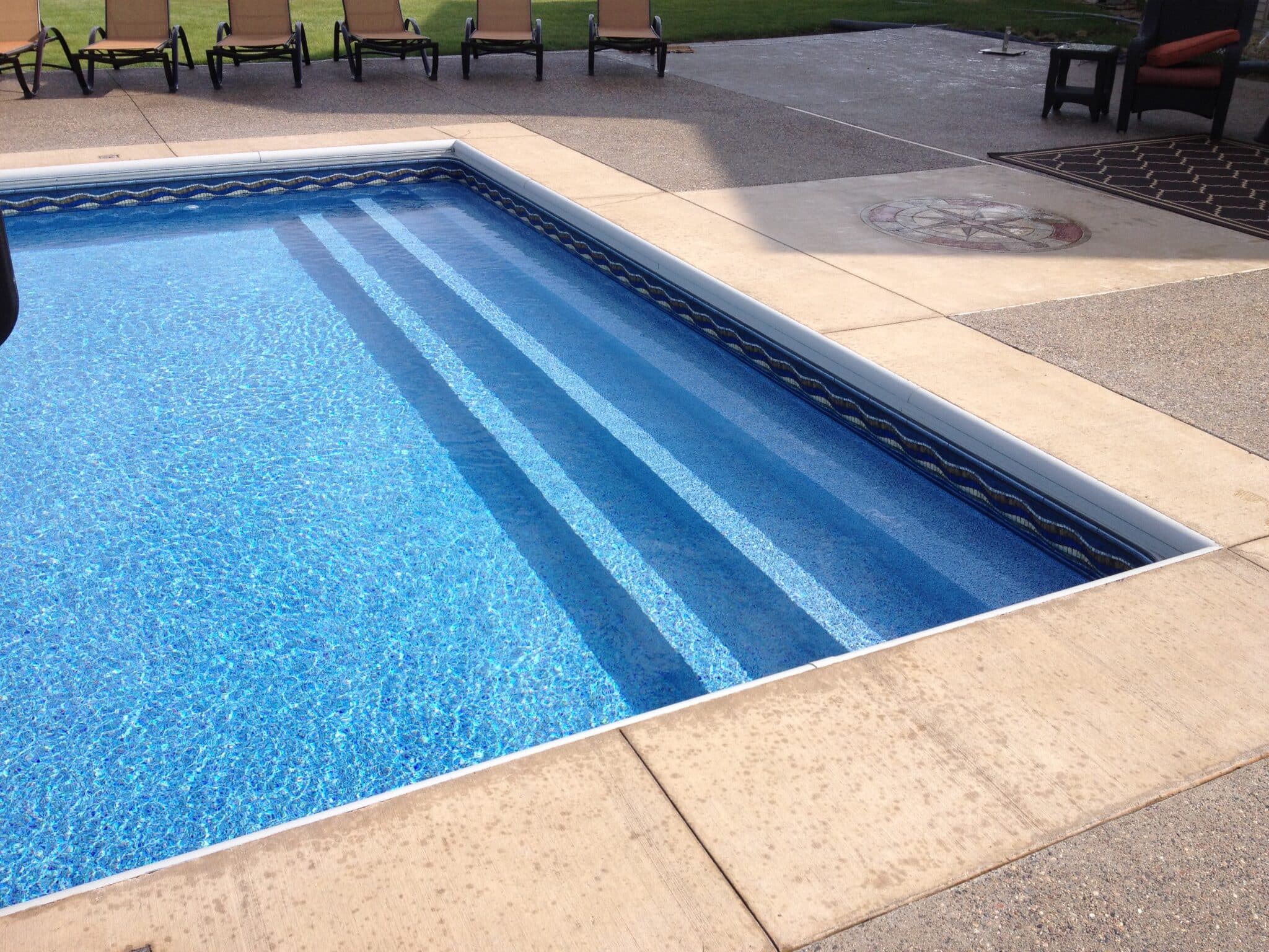 A sunny backyard pool area featuring clear blue water over a patterned vinyl liner, wide internal steps, and a tan concrete deck with lounge chairs in the background.